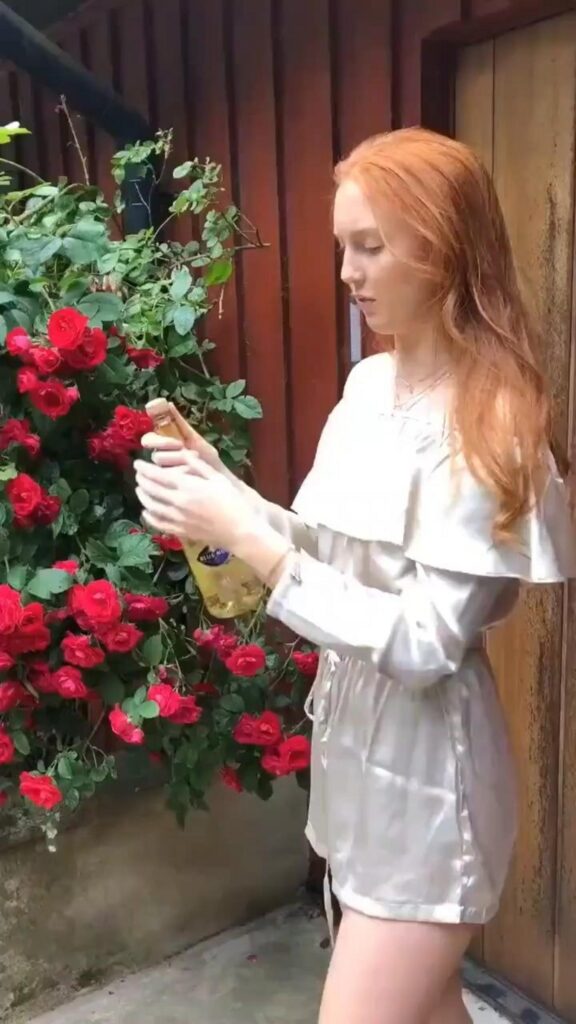 赤いバラの隣で瓶を開ける赤毛の女性